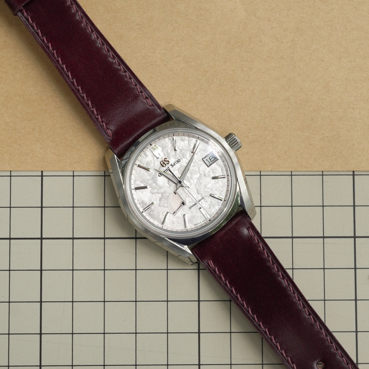 Burgundy shell cordovan slim strap on a Grand Seiko with a textured white dial, laid on a cutting mat