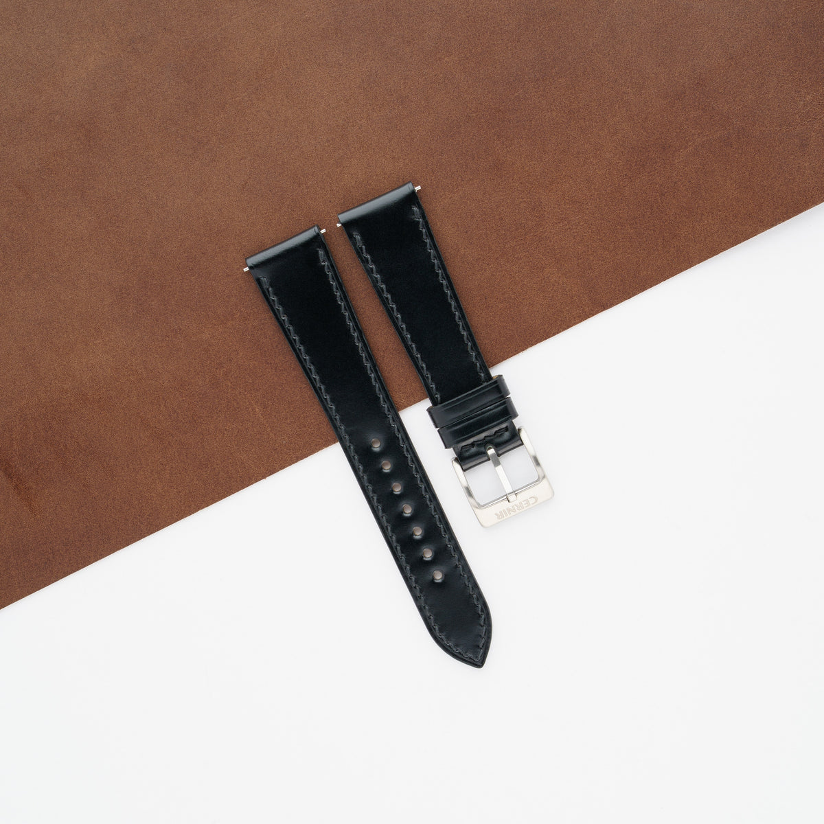 Glossy black shell cordovan slim watch strap laid across a brown leather sheet and white background