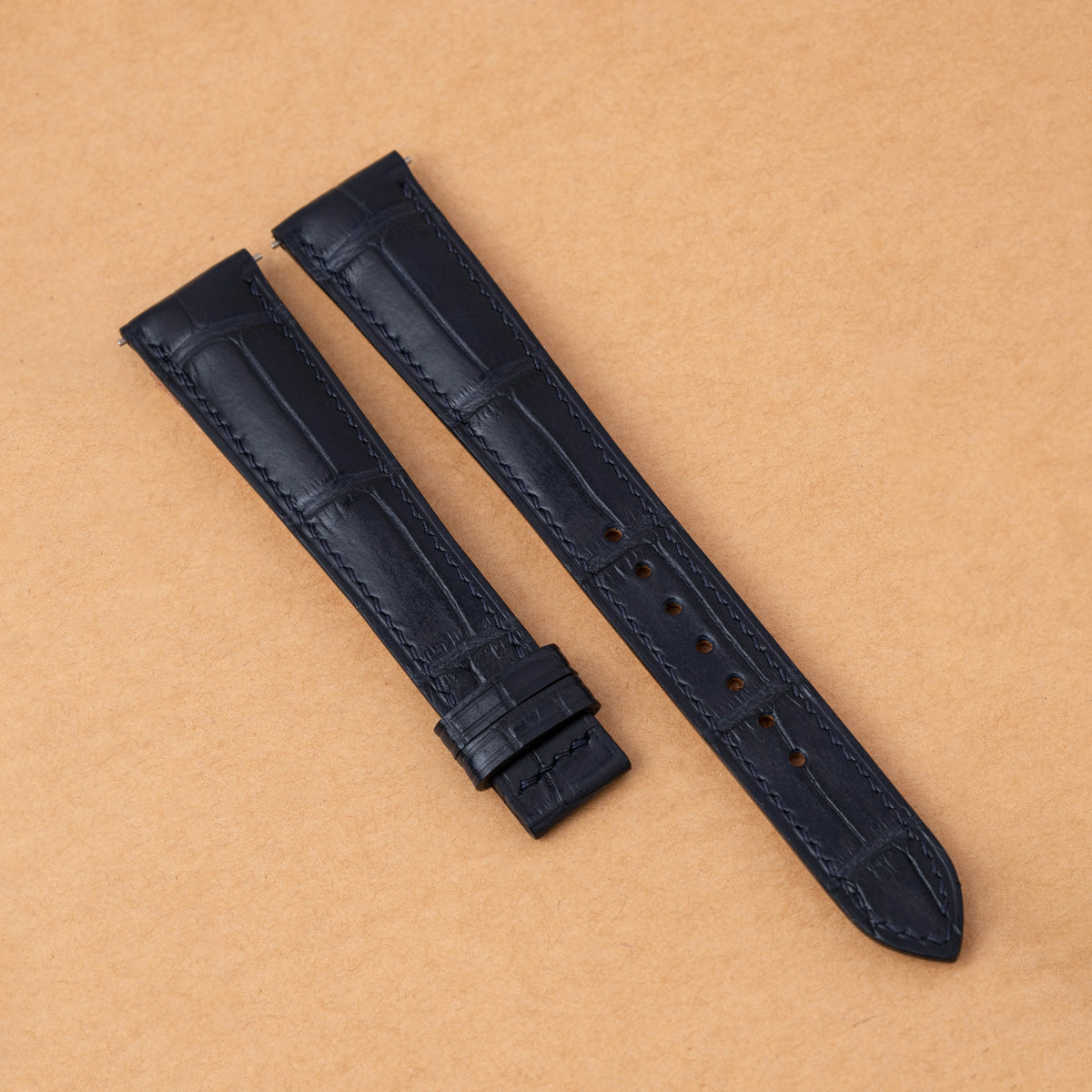 Navy alligator padded watch strap on a tan background, showing the square scales