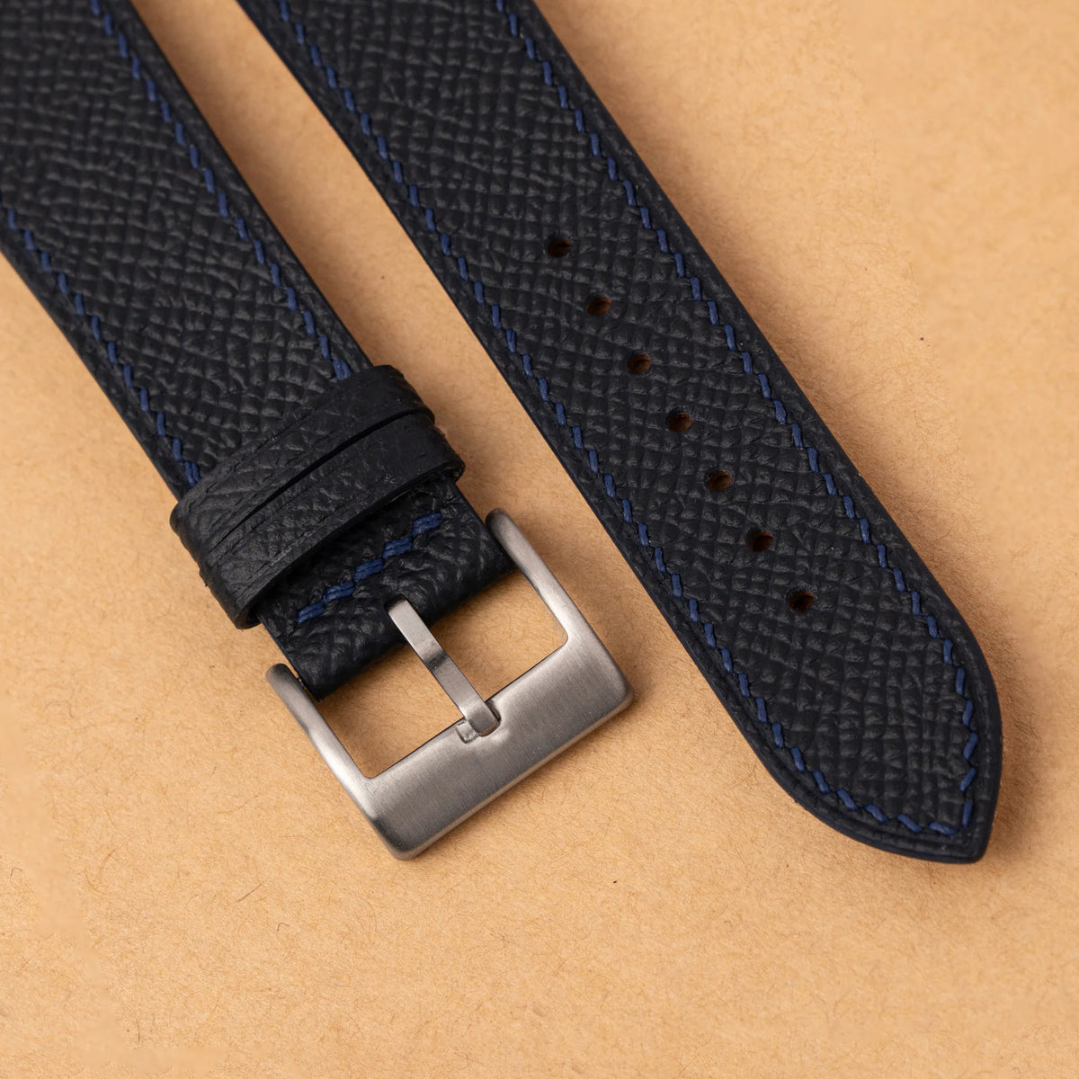 Close-up of the embossed Epsom grain and navy hand stitching beside the steel buckle of the black curved end strap