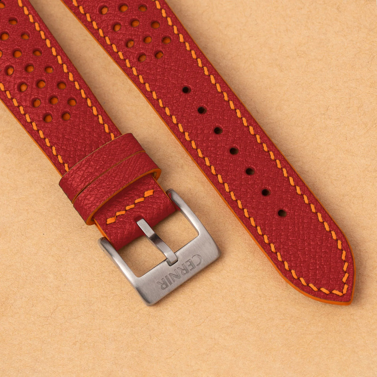 Close-up of round perforations over orange leather, the coral red Epsom grain and a silver Cernir buckle