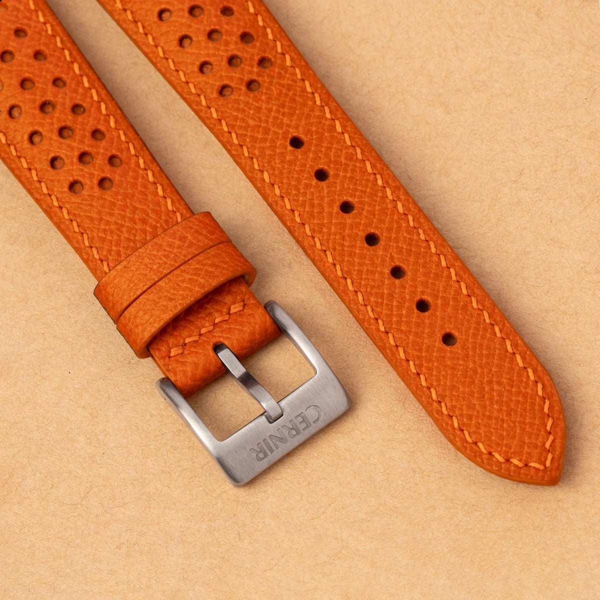 Close-up of round perforations over orange leather, the orange Epsom grain and a silver Cernir buckle