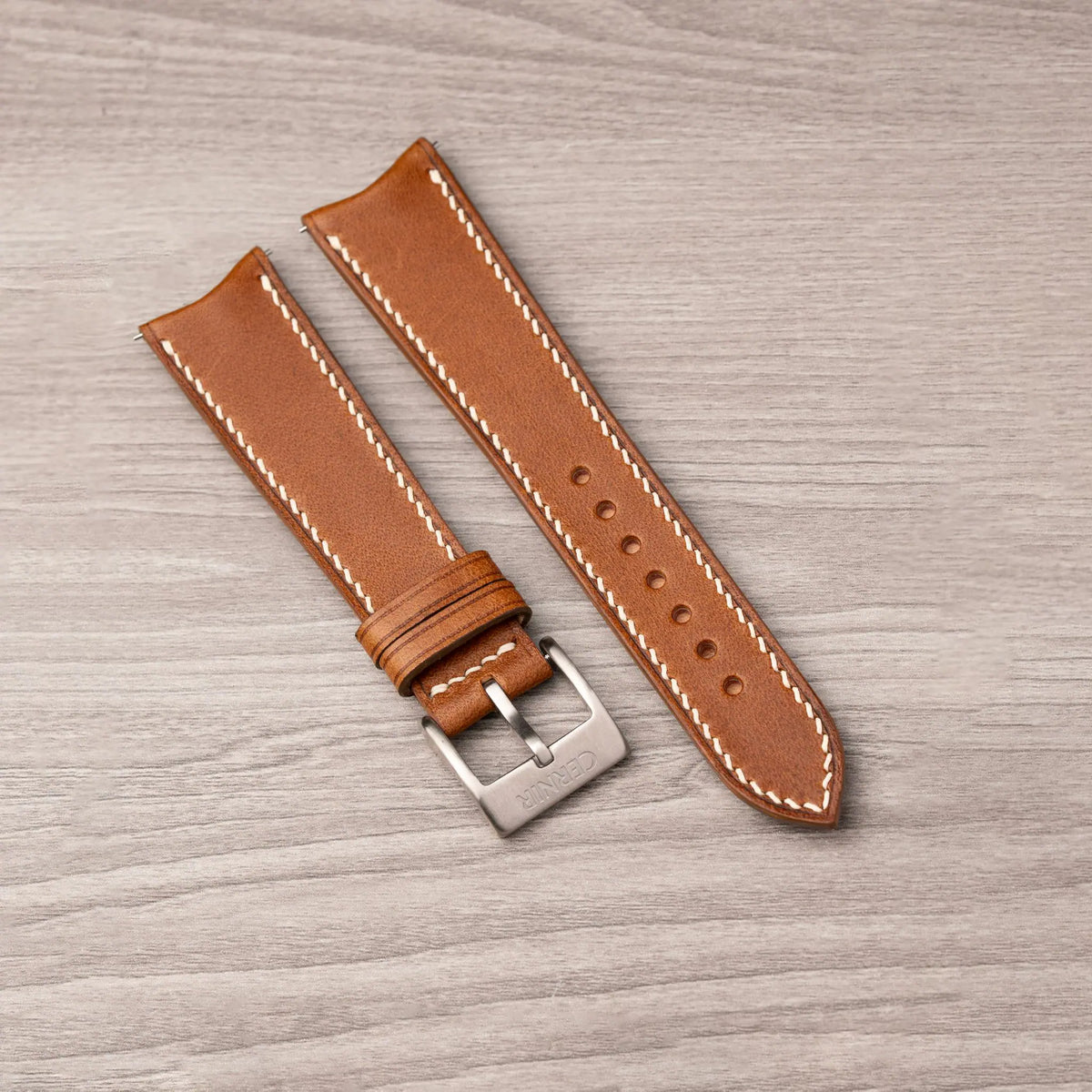 Tan Buttero curved end strap with cream stitching and a steel pin buckle on a wooden surface