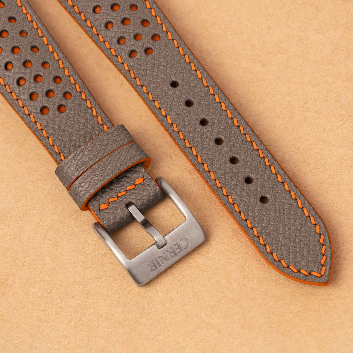 Close-up of round perforations over orange leather, the taupe Epsom grain and a silver Cernir buckle